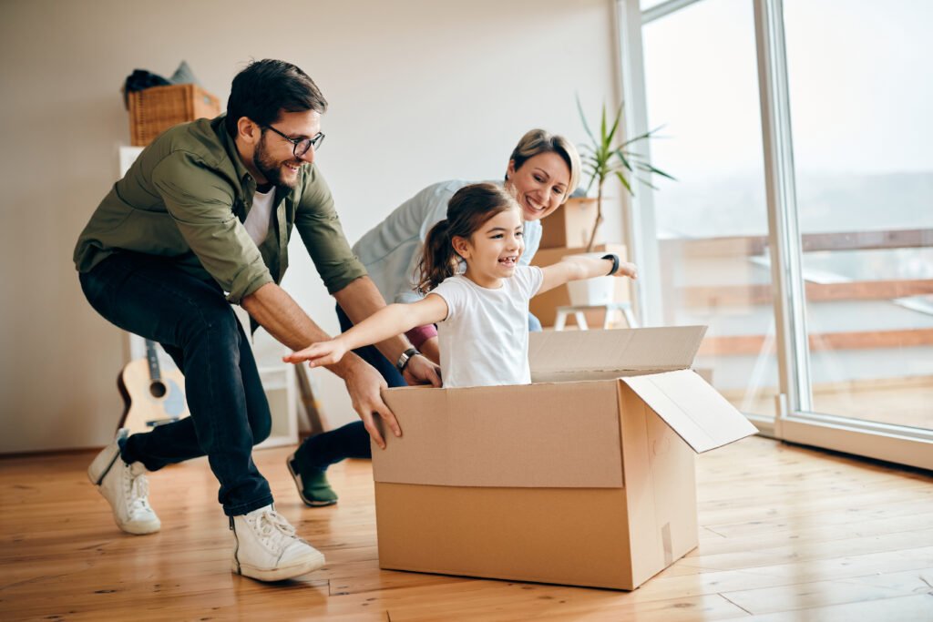 Happy daughter having fun with her parents while relocating in new apartment. Happy little girl with arms outstretched having fun being pushed by her parents in carton box at new home